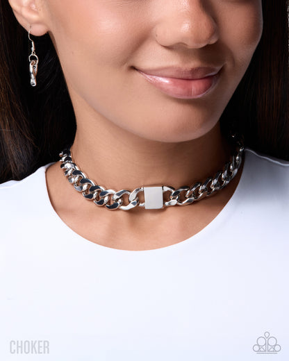 A close-up of a model wearing a chunky silver chain choker with a rectangular clasp, paired with a small silver drop earring and a white t-shirt.