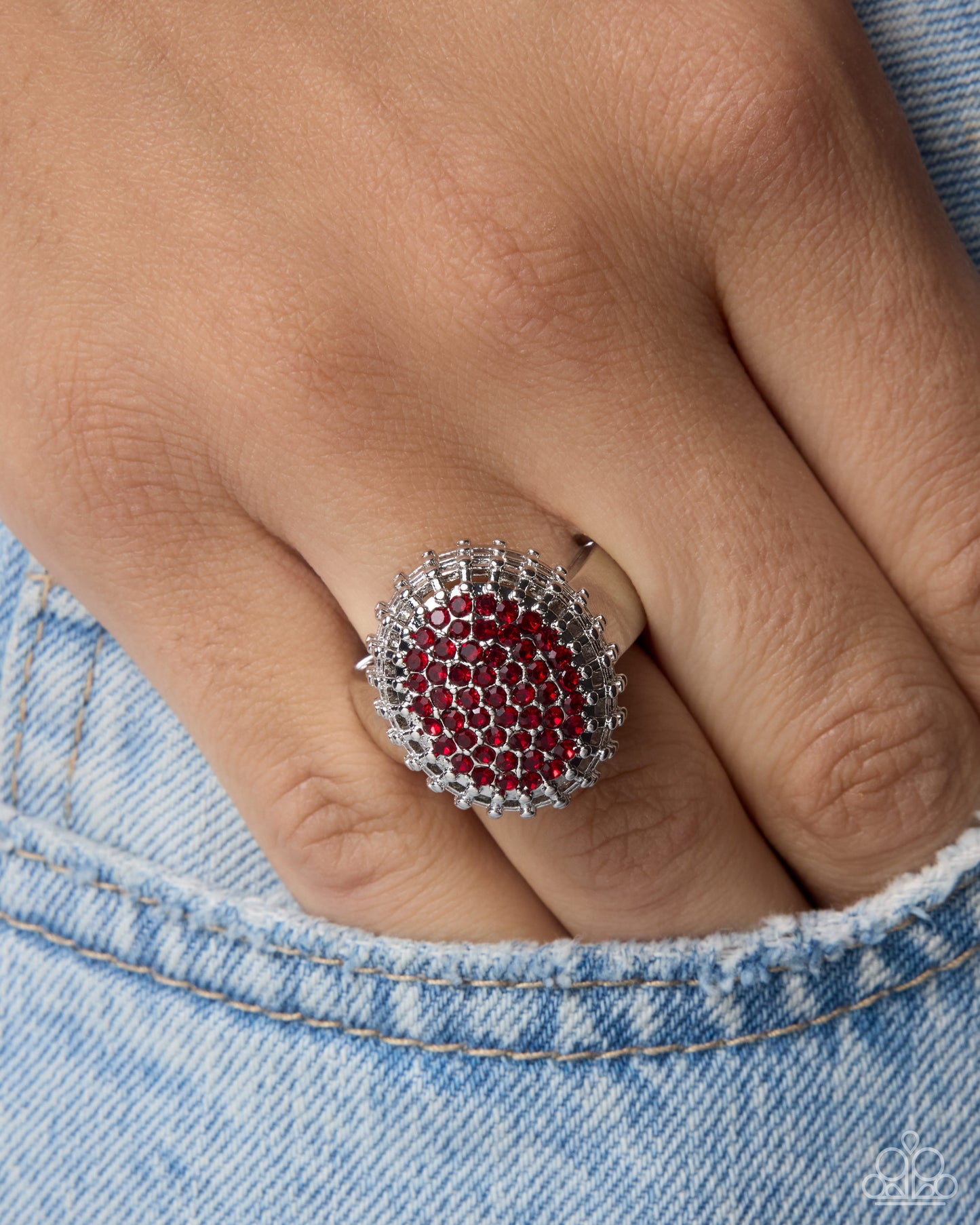 A close-up of a hand wearing a silver ring with a circular red gemstone center surrounded by clear crystals, resting against light blue denim.