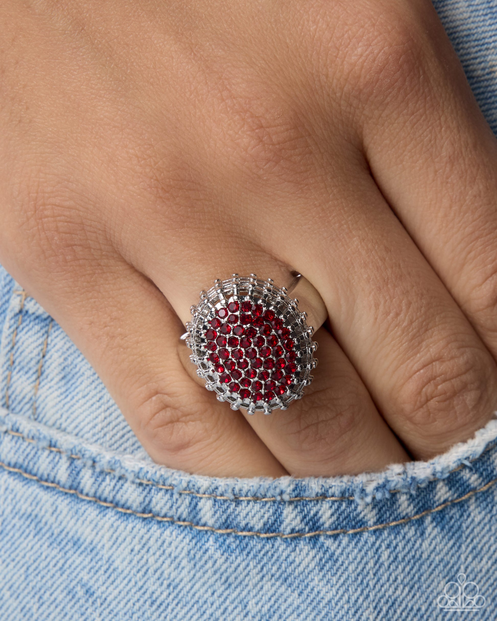 A close-up of a hand wearing a silver ring with a circular red gemstone center surrounded by clear crystals, resting against light blue denim.