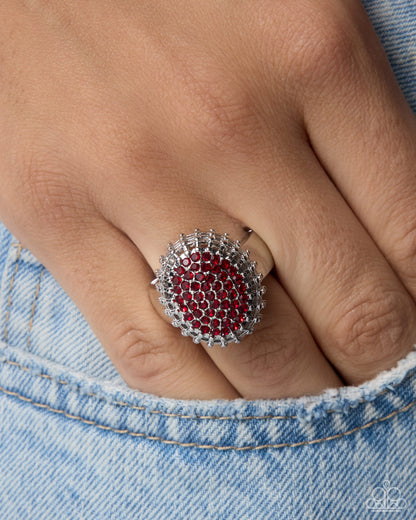 A close-up of a hand wearing a silver ring with a circular red gemstone center surrounded by clear crystals, resting against light blue denim.