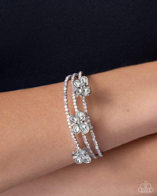 A close-up of a person's wrist wearing three stacked silver-toned bracelets, each decorated with a clear crystal flower motif, against a dark navy top background.