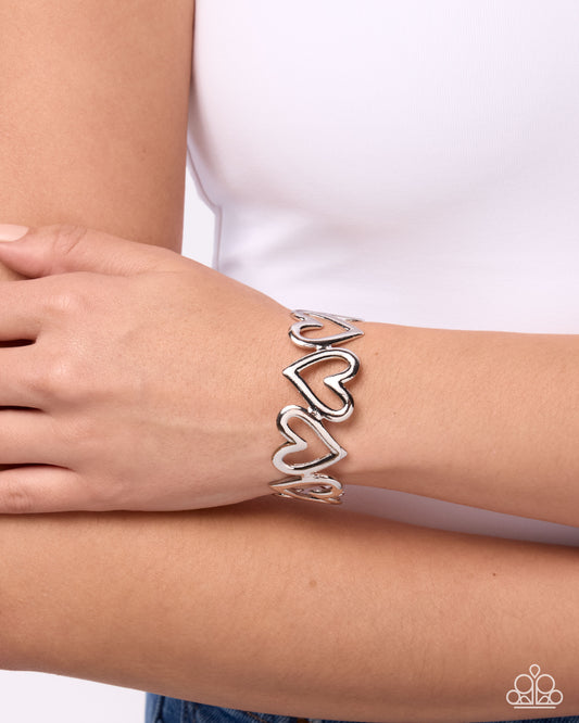 A silver cuff bracelet with three interlocking open heart designs, shown worn on a person's wrist against a white top background.