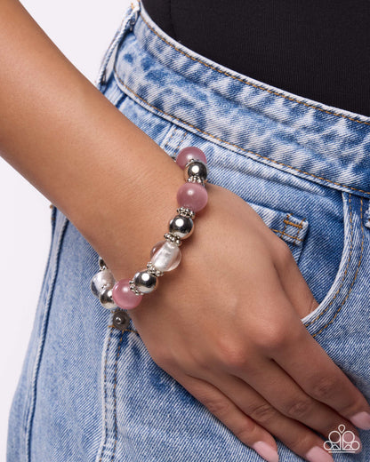 A close-up of a person's hand resting on their denim jeans, showcasing a beaded stretch bracelet with pink, white, and silver beads.