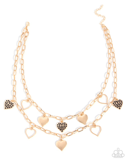 A double-layered gold-tone chain necklace with heart charms, featuring some heart charms encrusted with small black stones, displayed on a clean white background.