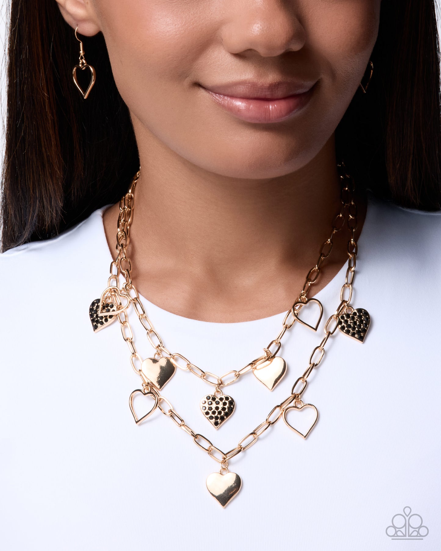 A close-up of a model wearing a gold multi-layered necklace with heart-shaped charms, including hollow and textured heart pendants, paired with matching heart earrings.