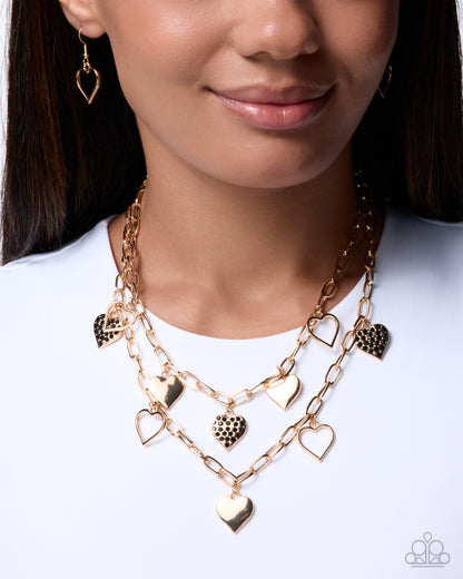 A close-up of a model wearing a gold multi-layered necklace with heart-shaped charms, including hollow and textured heart pendants, paired with matching heart earrings.