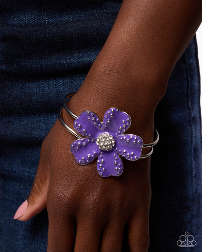 A close-up of a person's wrist wearing a silver-toned bangle bracelet with a large purple flower face, studded details, and a sparkling crystal-like center.