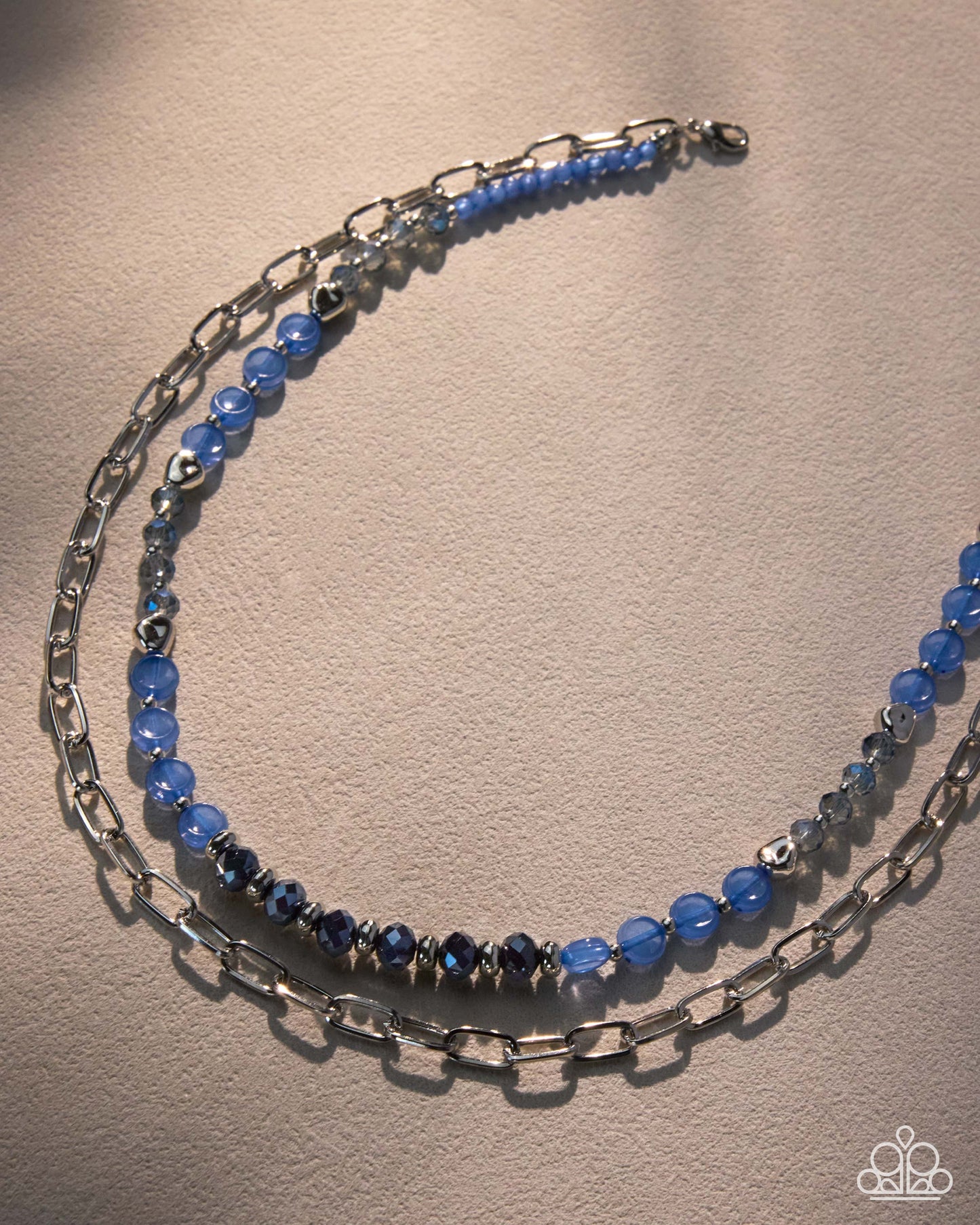 A double-strand beaded and chain necklace featuring light blue faceted beads, dark blue beads, and silver-toned metal links, displayed on a textured beige surface.