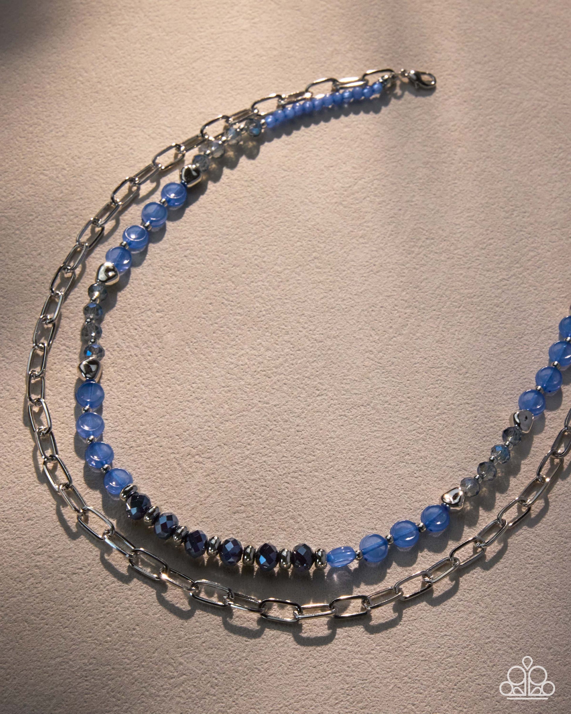 A double-strand beaded and chain necklace featuring light blue faceted beads, dark blue beads, and silver-toned metal links, displayed on a textured beige surface.