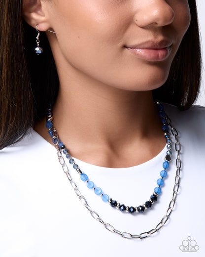 A close-up of a woman wearing a double-strand necklace from Paparazzi Jewelry. The necklace features a silver-tone paperclip chain layered with a beaded strand of light blue and dark blue faceted beads.