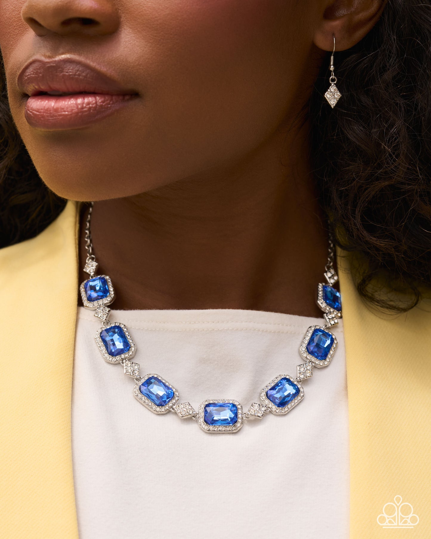 A close-up lifestyle shot of a woman wearing a silver-toned necklace with rectangular blue gemstones and a matching drop earring, styled with a white top and a yellow blazer.