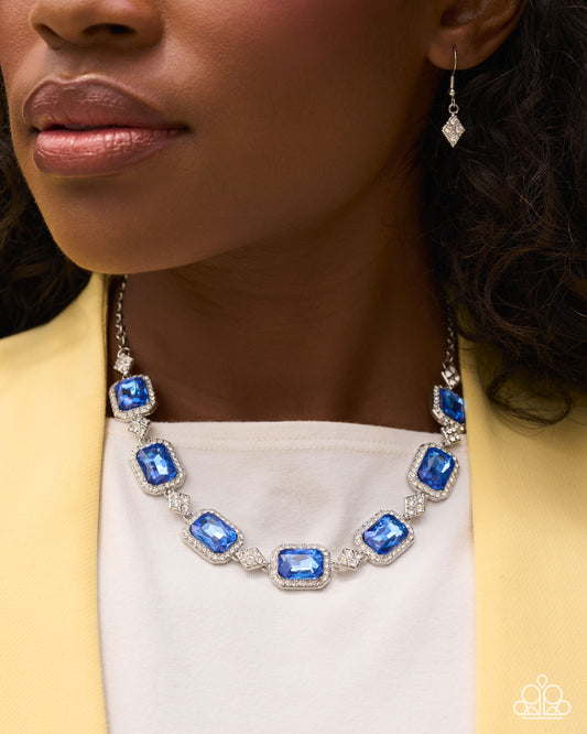 A close-up lifestyle shot of a woman wearing a silver-toned necklace with rectangular blue gemstones and a matching drop earring, styled with a white top and a yellow blazer.