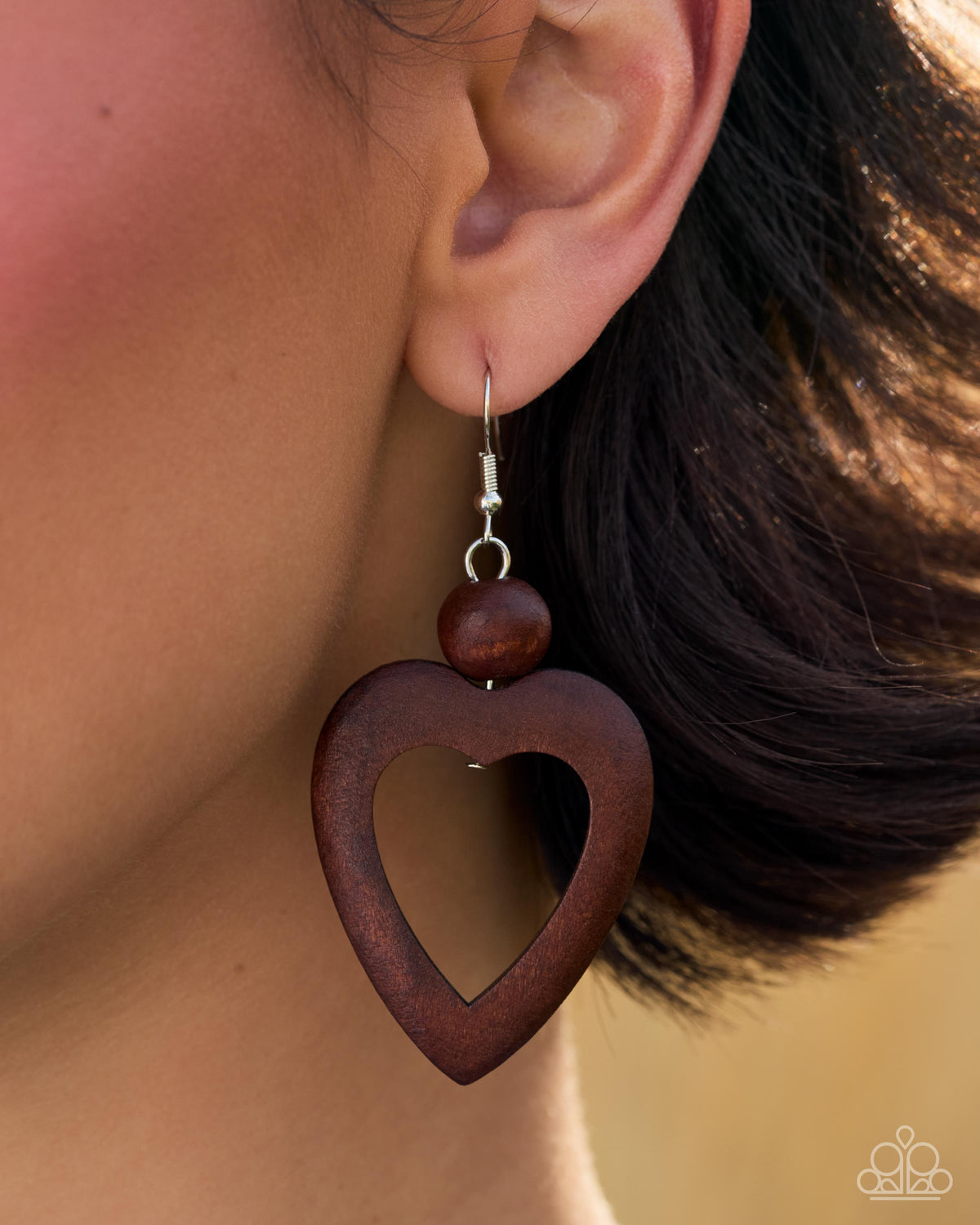 A close-up of a person's ear wearing a dark brown wooden heart-shaped dangle earring with a smaller round wooden bead and a silver fishhook wire above it.