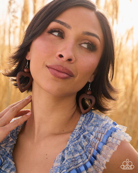 A woman wearing large heart-shaped wooden earrings with a natural wood grain finish, photographed outdoors against a warm, blurred background of dried grasses.