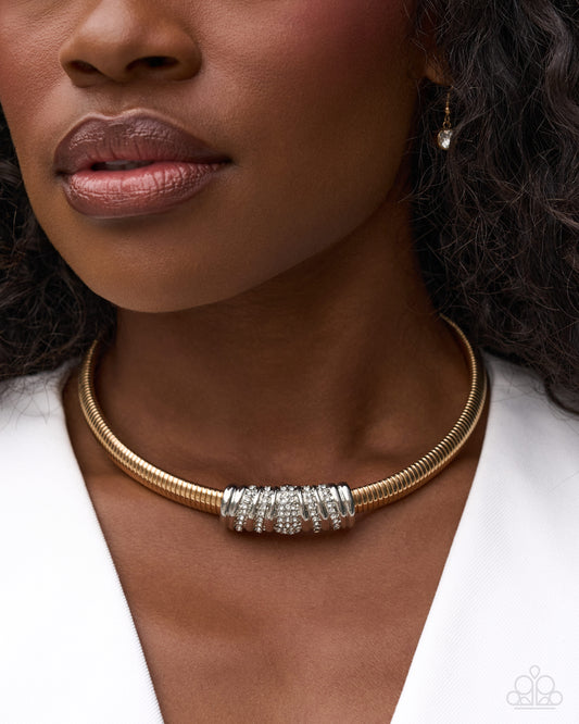 A close-up of a model wearing a gold snake chain choker necklace with a central silver-toned cylindrical cluster of clear stones, paired with a white top.
