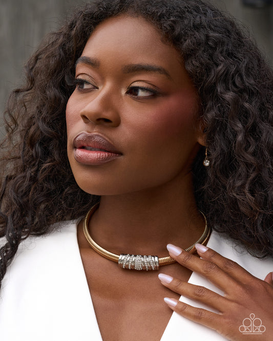 A close-up portrait of a woman with curly dark hair wearing a gold-toned bangle necklace with a textured silver-toned center, a matching drop earring, and a white top.