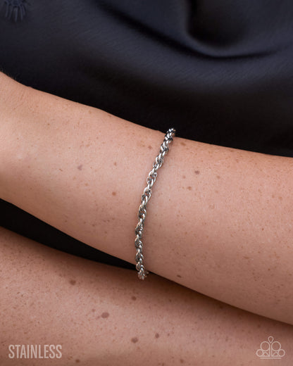 A close-up of a person's wrist wearing a thin twisted stainless steel chain bracelet, with a black top in the background and a 'STAINLESS' text watermark in the lower left corner.