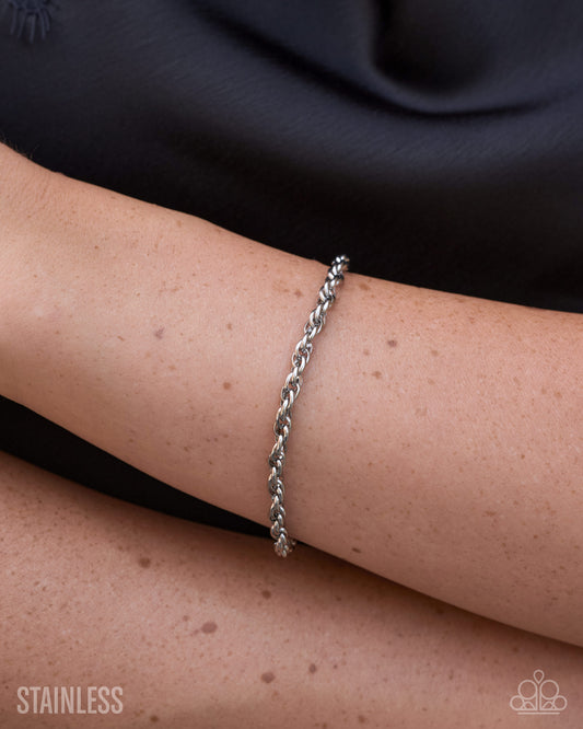 A close-up of a person's wrist wearing a thin twisted stainless steel chain bracelet, with a black top in the background and a 'STAINLESS' text watermark in the lower left corner.