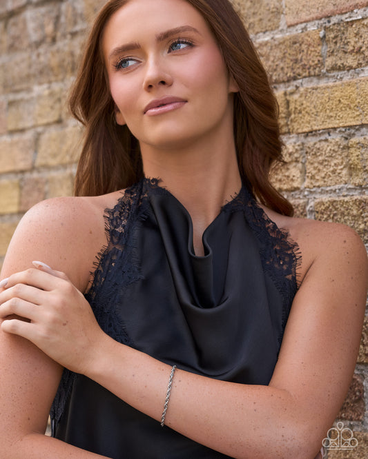 A woman with brown hair leans against a tan brick wall, wearing a black satin halter top with lace trim and a silver chain bracelet.