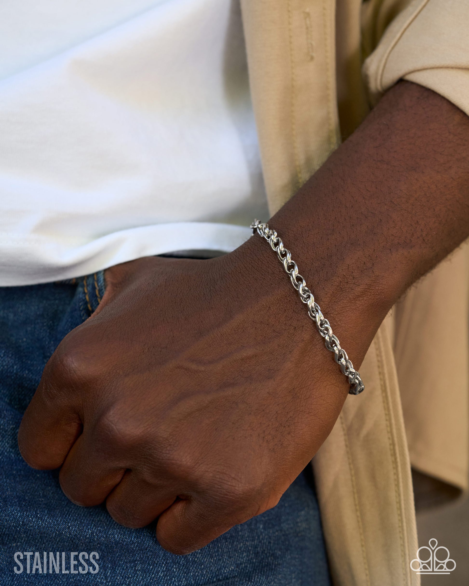A close-up of a person's wrist wearing a silver stainless steel curb chain bracelet, styled with a white t-shirt, beige overshirt, and blue jeans.
