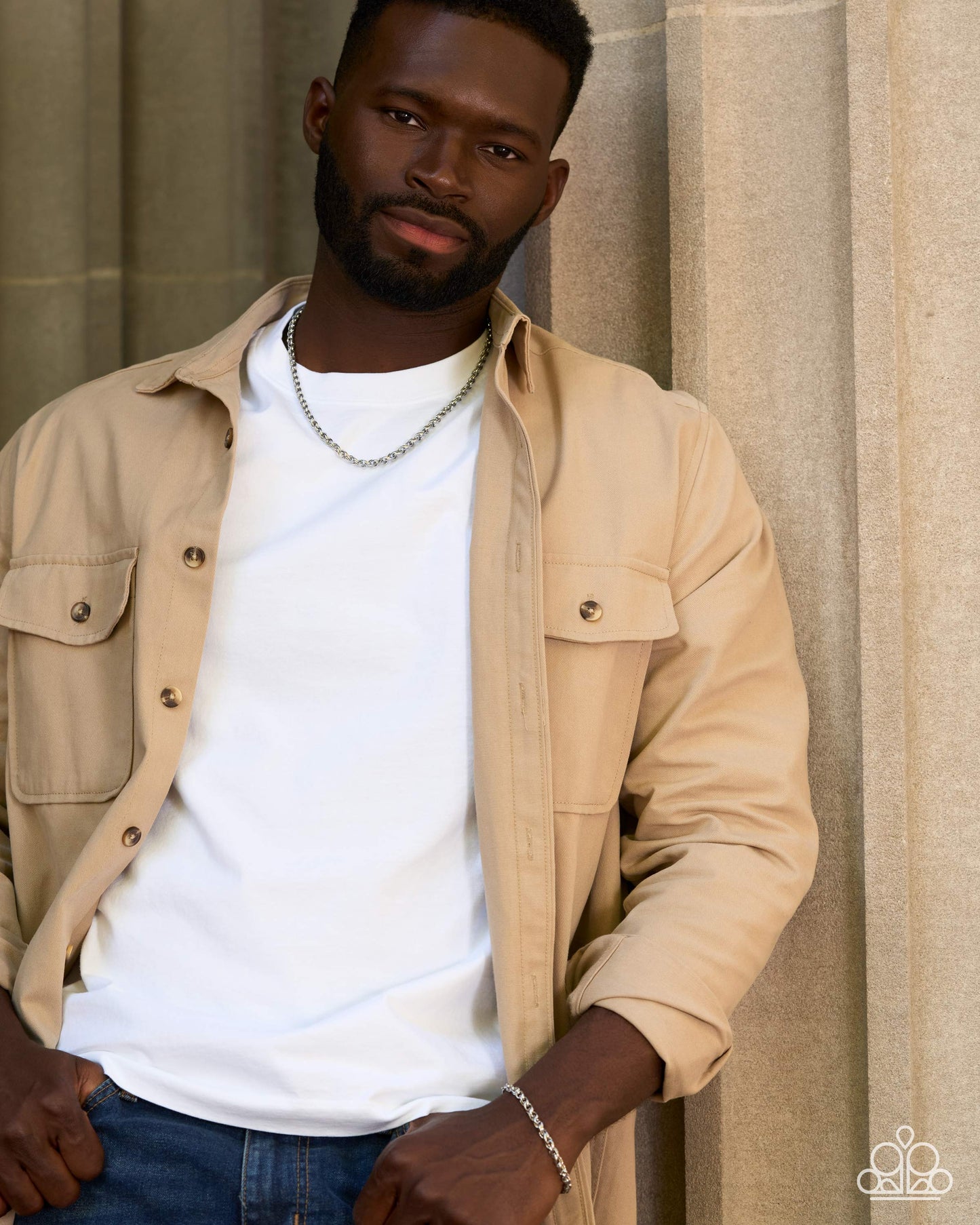 A man wearing a beige button-up overshirt over a white t-shirt and blue jeans, standing against a neutral beige stone wall background.
