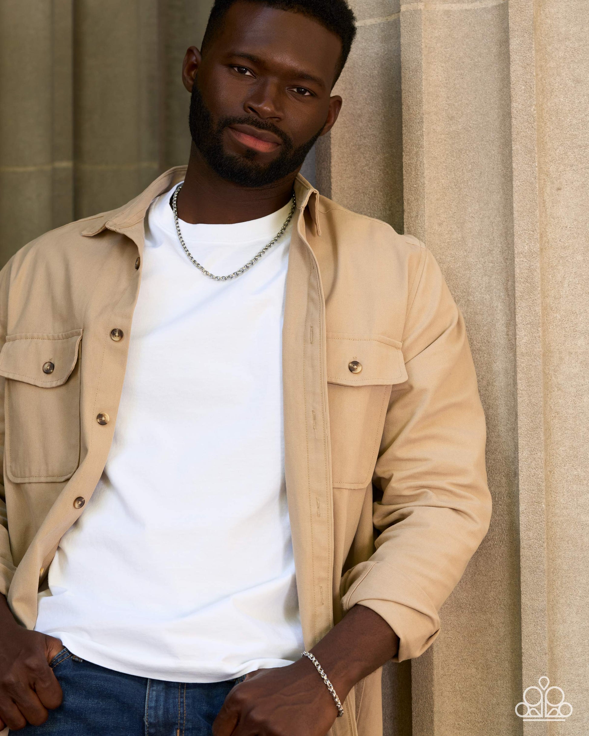 A man wearing a beige button-up overshirt over a white t-shirt and blue jeans, standing against a neutral beige stone wall background.