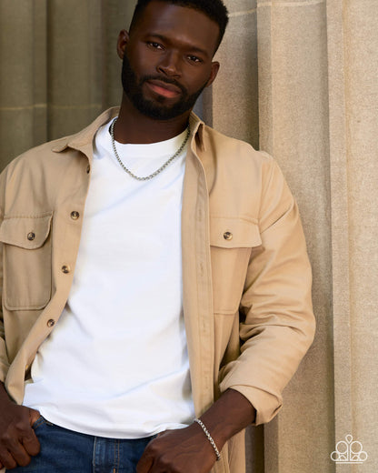A man wearing a beige button-up overshirt over a white t-shirt and blue jeans, standing against a neutral beige stone wall background.