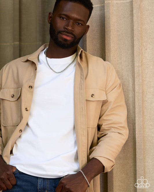 A man wearing a beige button-up overshirt over a white t-shirt and blue jeans, standing against a neutral beige stone wall background.