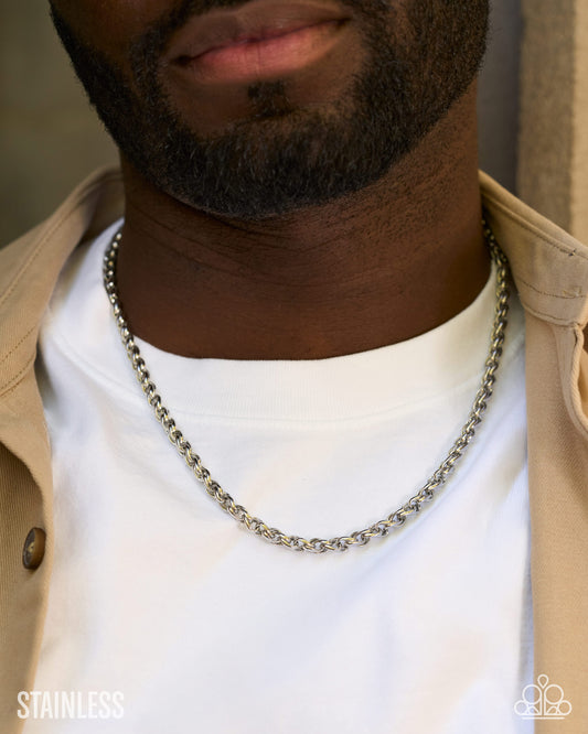 A close-up of a man wearing a silver stainless steel chain necklace over a white t-shirt and beige jacket.