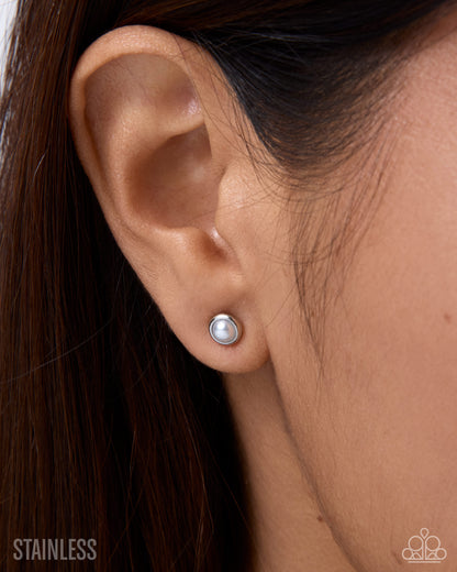 A close-up of a person's ear wearing a small round silver stud earring with a white pearl-like center. Dark brown hair frames the ear, and a silver 'STAINLESS' text overlay is visible in the bottom left corner.