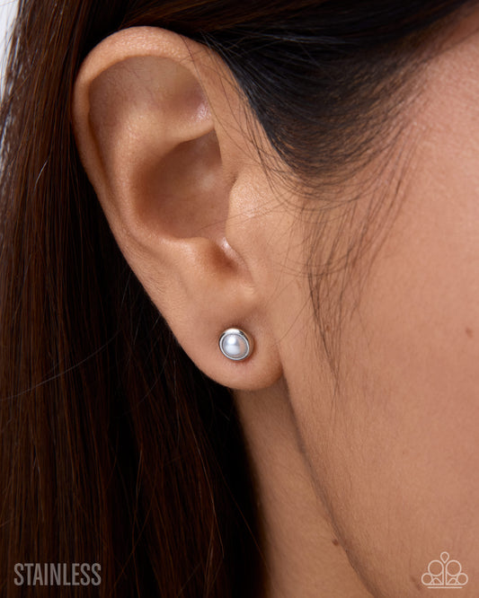 A close-up of a person's ear wearing a small round silver stud earring with a white pearl-like center. Dark brown hair frames the ear, and a silver 'STAINLESS' text overlay is visible in the bottom left corner.