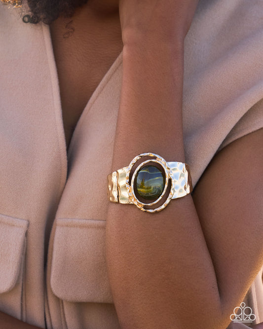 A close-up of a model's arm wearing a chunky gold cuff bracelet with a large oval green cabochon stone.