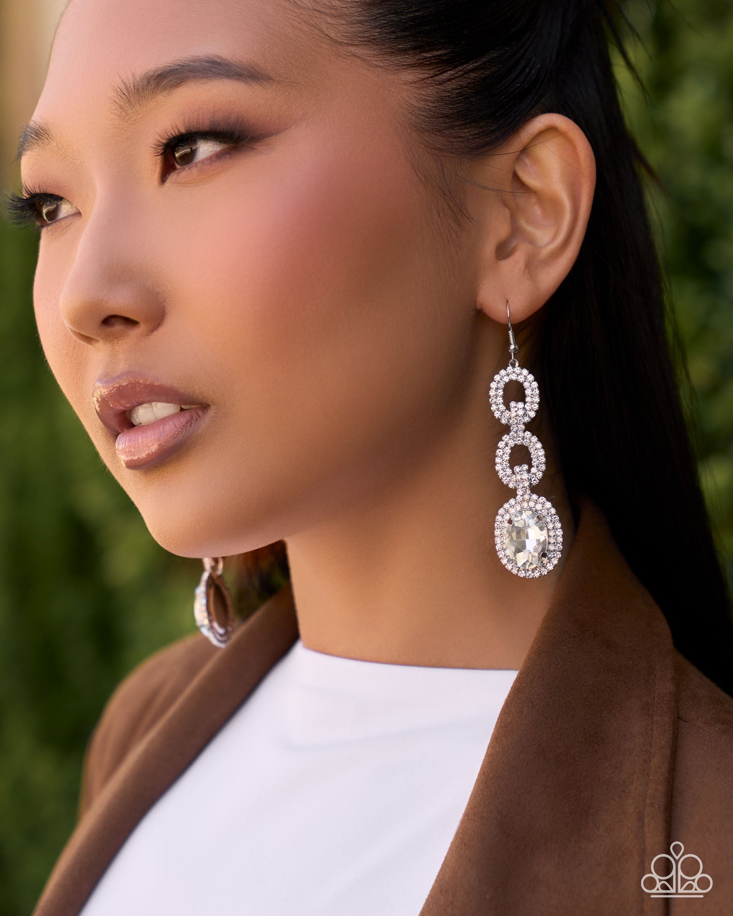 A close-up side profile of a woman wearing a long, three-link drop earring with sparkling clear stones in a silver-tone setting, paired with a brown blazer and white top against a soft green blurred background.