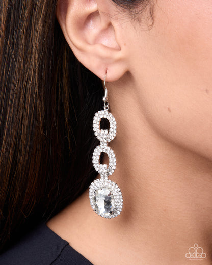 A close-up of a model's ear and neck wearing a long, dangling statement earring. The earring features three stacked oval links, each encrusted with small clear stones, with the largest oval link at the bottom featuring a larger clear marquise-cut center stone in a halo setting.