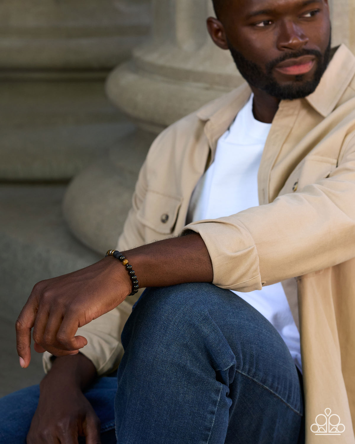 A man wearing a black beaded bracelet with gold accents, sitting outdoors against a stone column. He is dressed in a beige overshirt over a white t-shirt and blue jeans.
