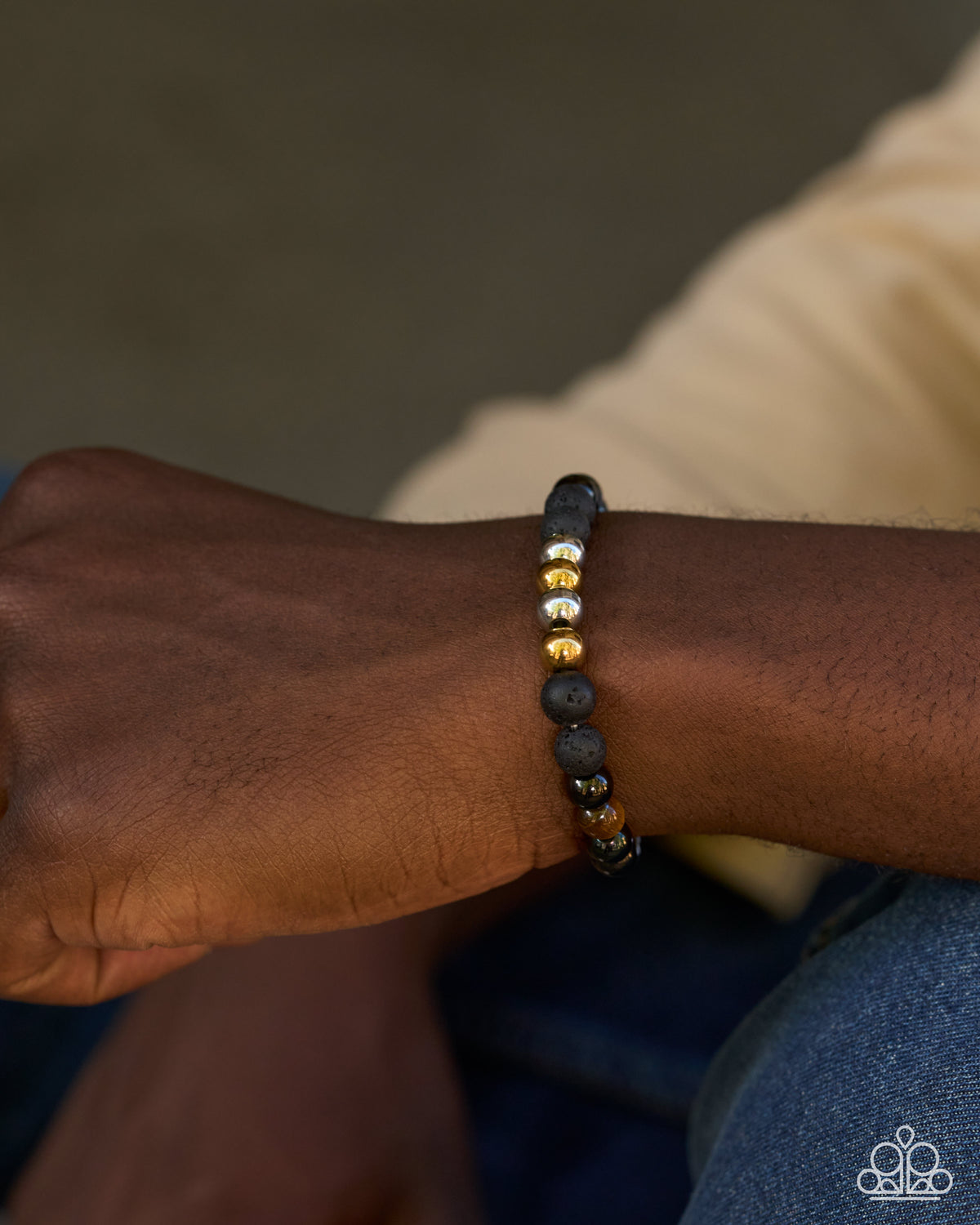 A close-up of a person's wrist wearing a beaded bracelet with dark gray and metallic gold beads, set against a soft-focus background of beige clothing and blue jeans.