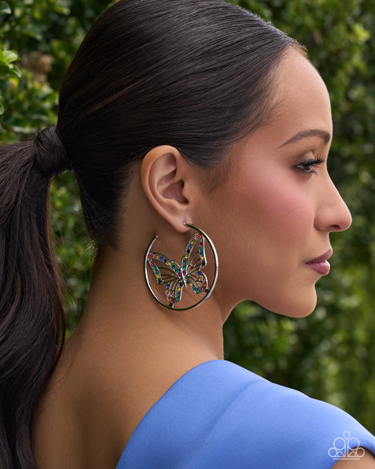 A woman in profile wearing large silver hoop earrings with a colorful butterfly inlay, set against a blurred green foliage background.