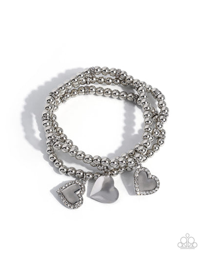 A set of three silver beaded stretch bracelets with heart-shaped charms, including one smooth silver heart and two heart charms encrusted with small clear rhinestones, on a white background.