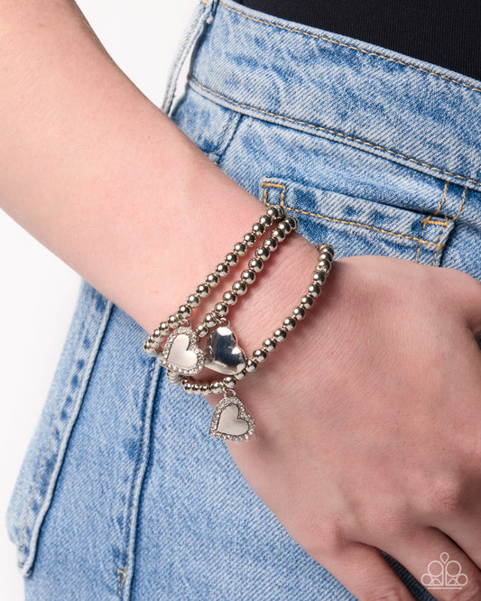 A close-up of a person's wrist wearing three beaded wrap bracelets with silver heart charms, paired with light blue denim jeans.