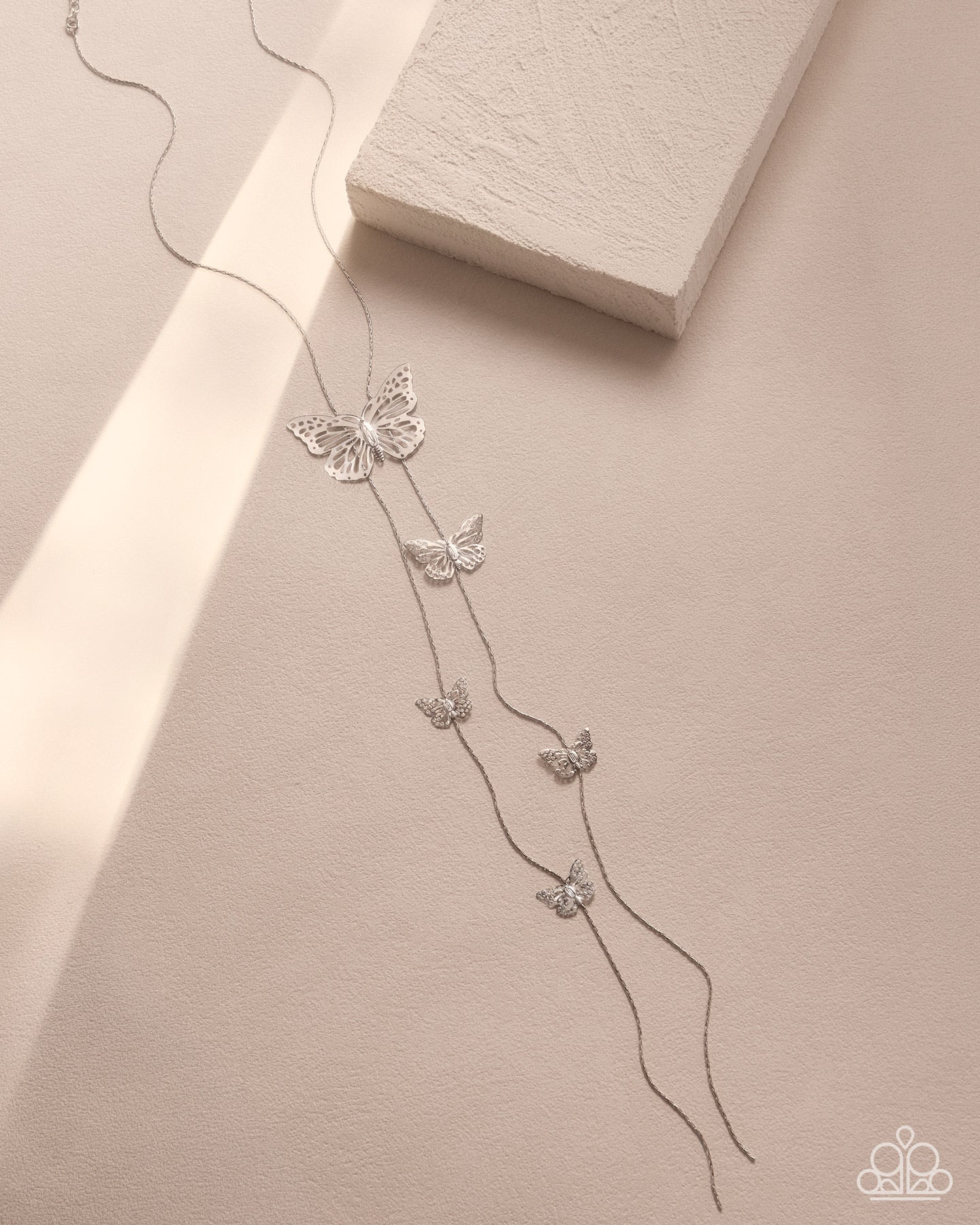 A delicate silver-toned necklace with five small butterfly charms arranged along a long chain, displayed on a warm beige surface next to a textured white block.