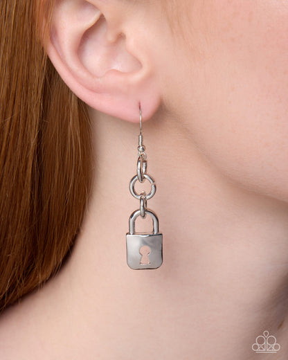 A close-up of a person's ear wearing a silver dangle earring shaped like a small padlock, with a keyhole cutout on the front.