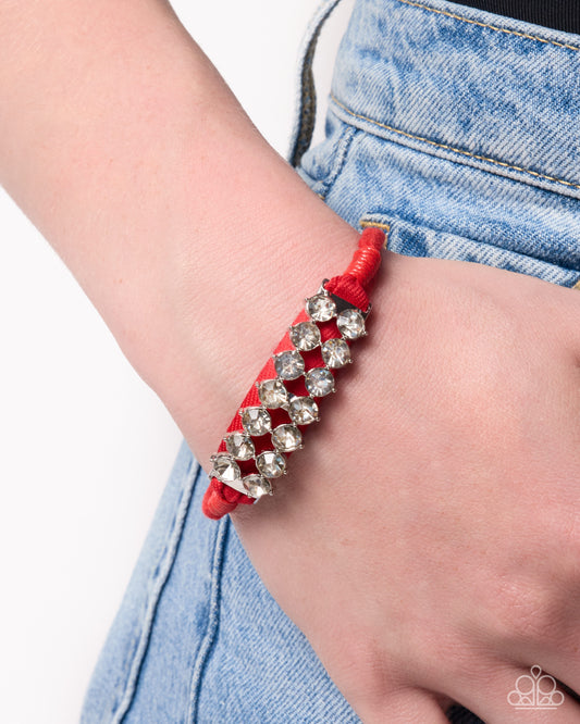 A close-up of a person's wrist wearing a red cord bracelet adorned with silver-toned rhinestone studs, paired with light blue denim jeans against a white background.
