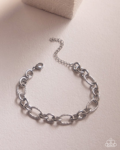 A silver chain link bracelet with a lobster clasp and extender chain, displayed on a textured beige surface.