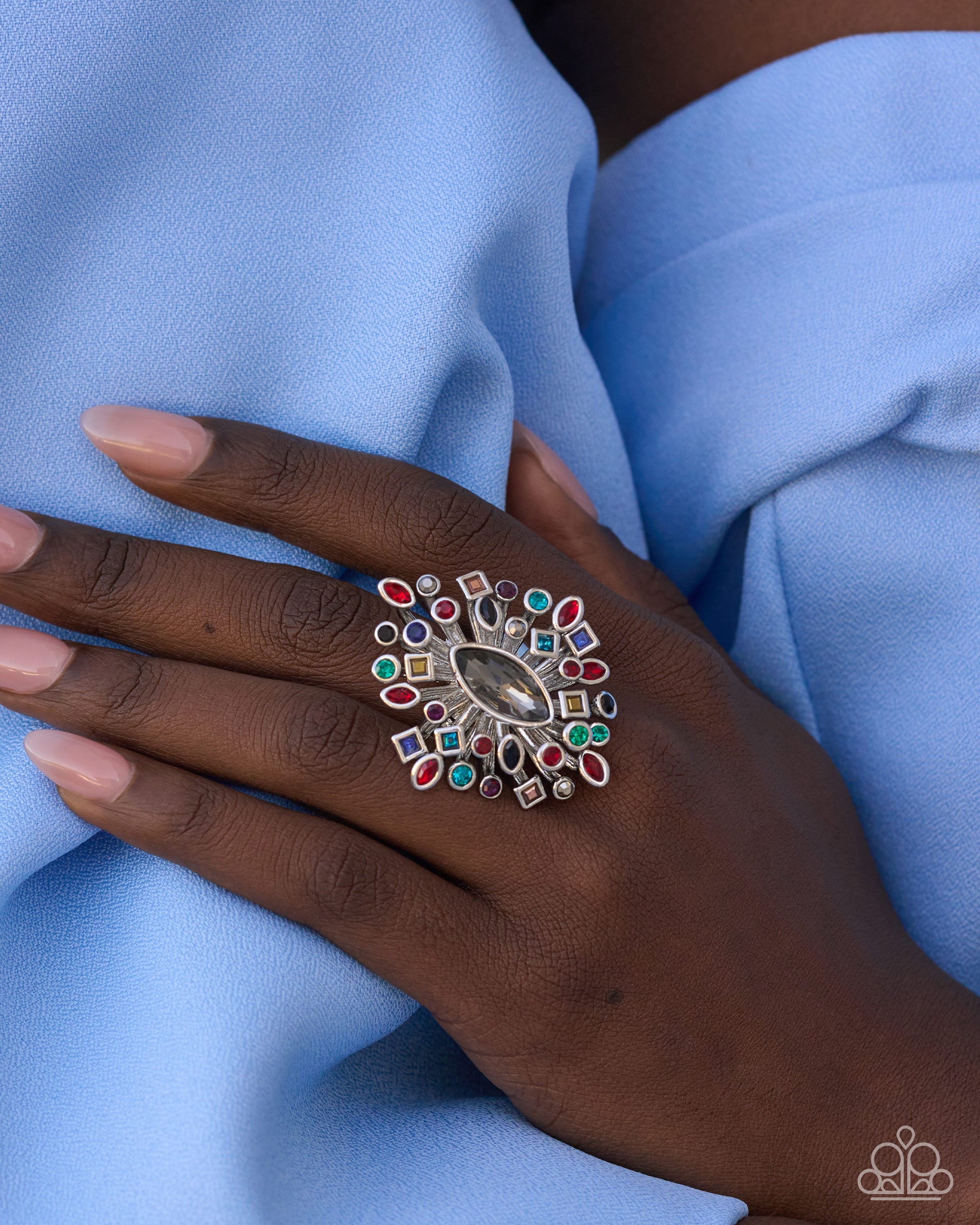 A close-up of a hand wearing a large, ornate silver ring decorated with multiple colorful gemstones, resting on light blue fabric.