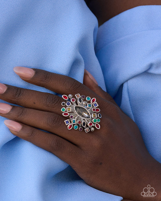 A close-up of a hand wearing a large, ornate silver ring decorated with multiple colorful gemstones, resting on light blue fabric.
