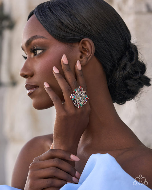 A close-up side profile of a woman with dark hair in an updo, wearing a large, ornate silver ring embellished with red and green stones. She is wearing a light blue strapless dress, and the background is a softly blurred beige stone wall.