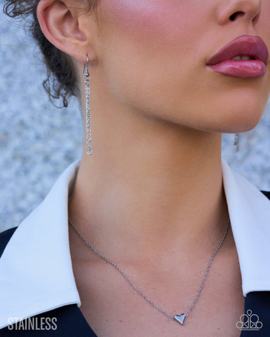 Close-up of a woman wearing a silver necklace with a small triangular pendant and matching long drop earrings, shown against a blurred outdoor background. The word 'STAINLESS' appears in the lower left corner.