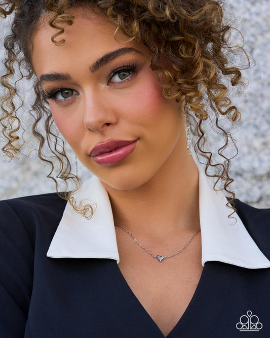 A close-up portrait of a woman with curly brown hair wearing a silver necklace with a tiny triangular pendant, paired with a white collar and dark navy top. The image has a small logo in the bottom right corner.