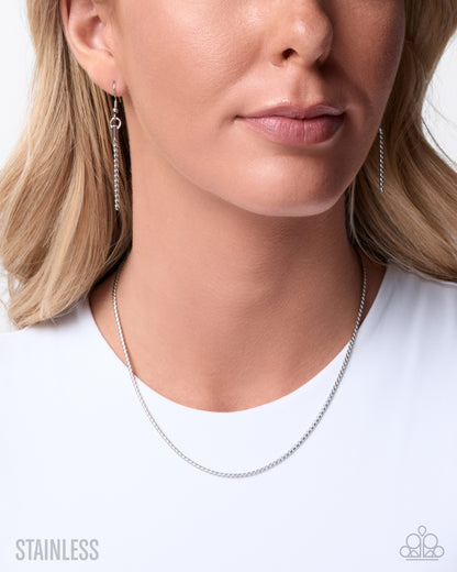 A close-up of a woman wearing a delicate stainless steel chain necklace and matching drop earrings, with a white t-shirt against a clean white background. Text overlays read 'STAINLESS' on the bottom left and a logo on the bottom right.