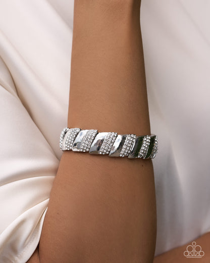 A close-up of a person's wrist wearing a chunky silver cuff bracelet embellished with rows of clear crystals, set against a soft cream-colored satin background.